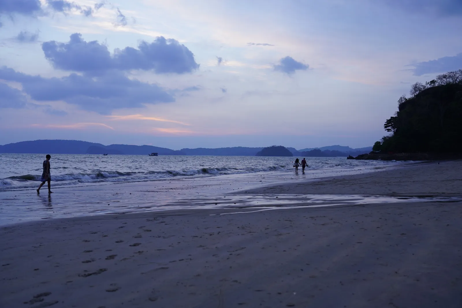 A praia de Ao Nang ao crepúsculo com água calma, algumas pessoas na areia e um promontório à direita