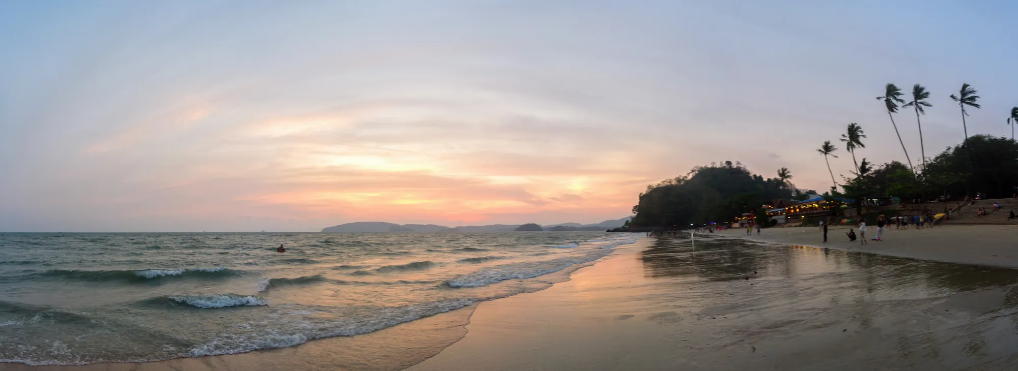 A praia de Ao Nang ao pôr do sol, com um céu cor-de-rosa, pequenas ondas na areia e palmeiras na margem oposta