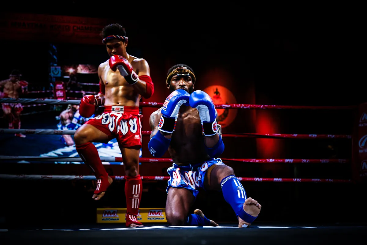 Dva bojovníci Muay Thai předvádějí ram muay v ringu pod scénickým osvětlením, jeden s vyzdviženým kolenem a jeden klečící v modrých rukavicích