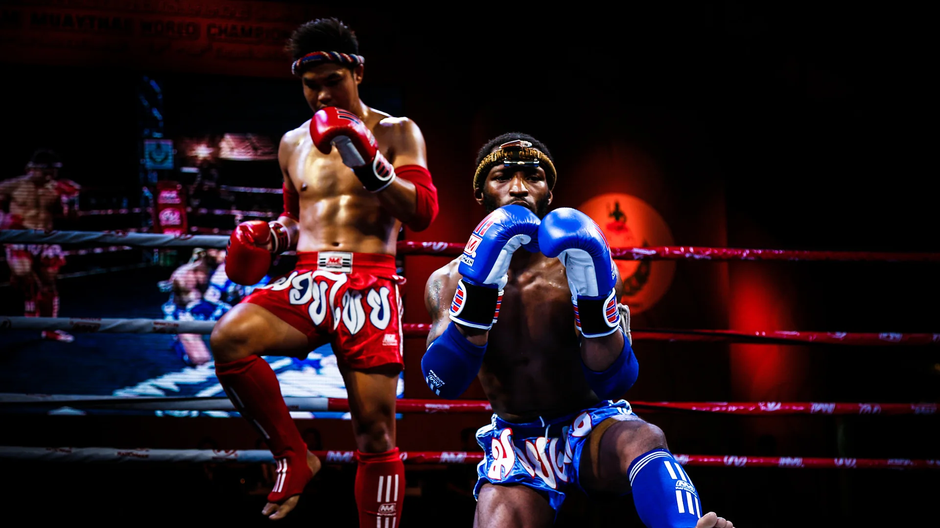 Dois lutadores de Muay Thai a executar o ram muay num ringue sob as luzes do palco, um de pé com o joelho levantado e luvas vermelhas e outro ajoelhado com luvas azuis
